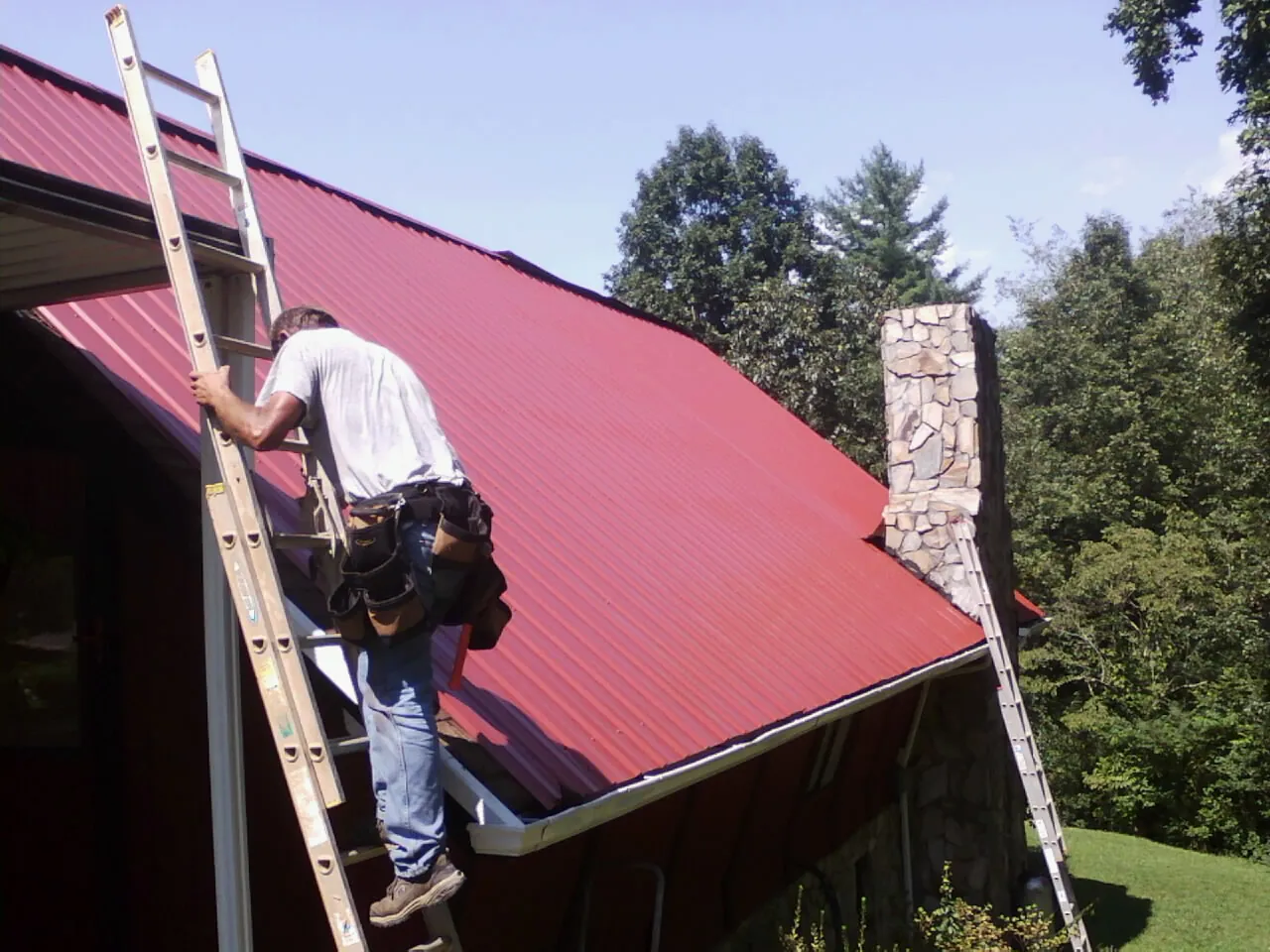Roofer on a ladder beside a red metal roof and stone chimney — Soffit & Fascia Repair by Roofing Brothers in Jonesville